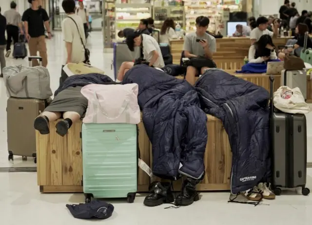 寝袋を使いベンチの上で寝る人たち。スーツケースが周りおいてある。後方のベンチには大勢が座っている