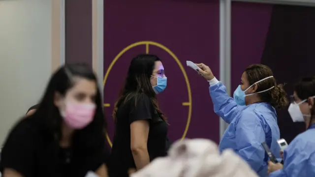 Duas profissionais e duas passageiras durante triagem em aeroporto; todas aparecem de máscara e uma funcionária estende equipamento portátil para verificar temperatura