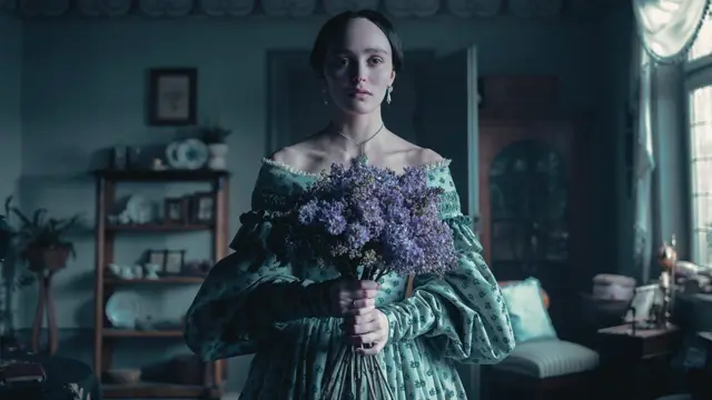 A atriz Lily-Rose Depp com vestido de época e segurando um buquê de flores roxas em cena do filme Nosferatu