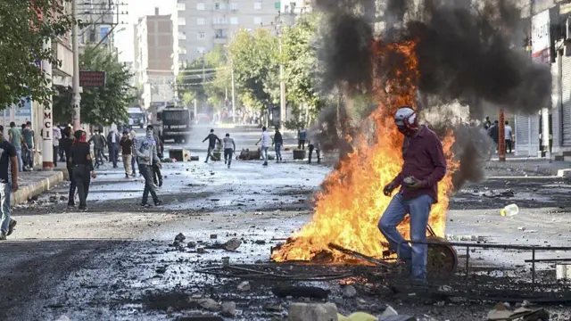 2014'te, Diyarbakır'daki 6-8 Ekim olaylarında yaşanan bir sokak çatışması. Yerde eşyalar yanıyor. Yüzleri kapalı eylemciler ellerinde taşla yürüyor.