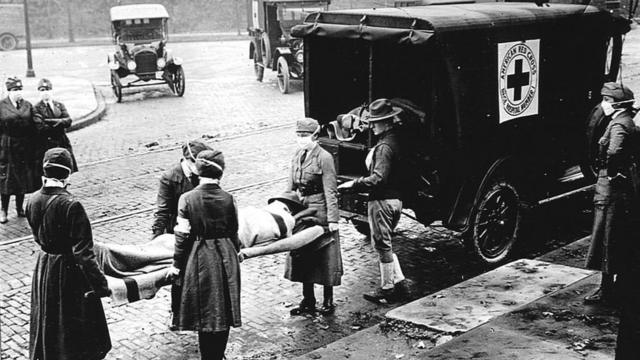 Membros do Corpo de Bombeiros da Cruz Vermelha, todos usando máscaras contra a propagação da epidemia de gripe, transportam um paciente em uma maca para a ambulância, em Saint Louis, Missouri, em outubro de 1918