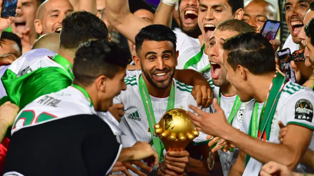 Le capitaine algérien Riyad Mahrez, vêtu d'un maillot blanc et arborant la médaille de vainqueur sur un ruban vert, sourit en brandissant le trophée de la Coupe d'Afrique des Nations, entouré de ses coéquipiers en liesse qui célèbrent la victoire avec lui.