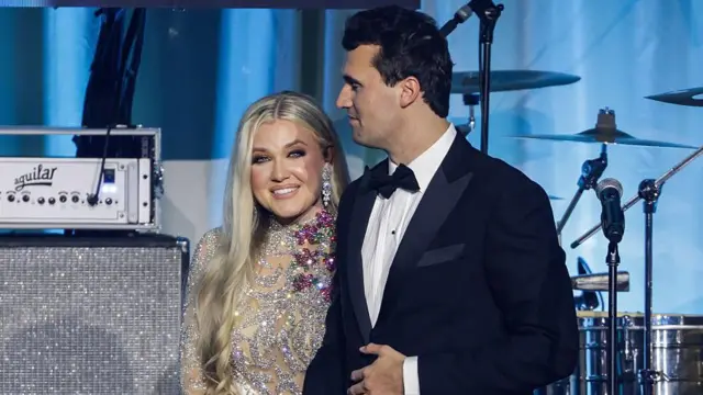 Erika e Charlie em pé no palco, sorrindo, com roupas de gala em frente a instrumentos musicais