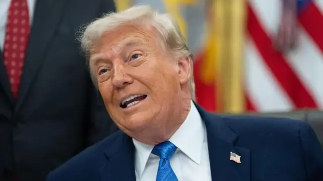 大統領執務室の机に向かって座り、記者団に話すトランプ氏。紺色のスーツに青いネクタイを締め、星条旗のピンバッジを襟に着けている