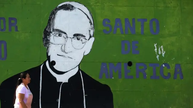 Imagem mostra mulher passandobetánofrente à parede pintada com imagem do monsenhor Oscar Arnulfo Romero, o novo santo da Igreja