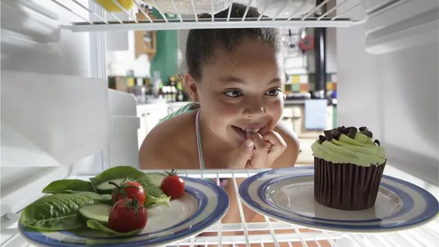 Menina olhando dentro da geladeira, onde está um pratocng casinosalada e um prato com cupcake