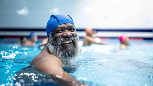 An elderly man smiling in a swimming pool smiling while other people swim and stand in the pool behind him
