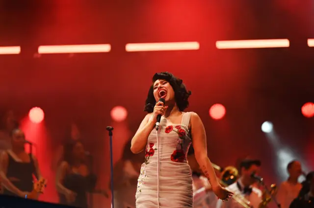 Raye sings into a microphone on a red-lit stage, wearing a nude dress with red flowers on.