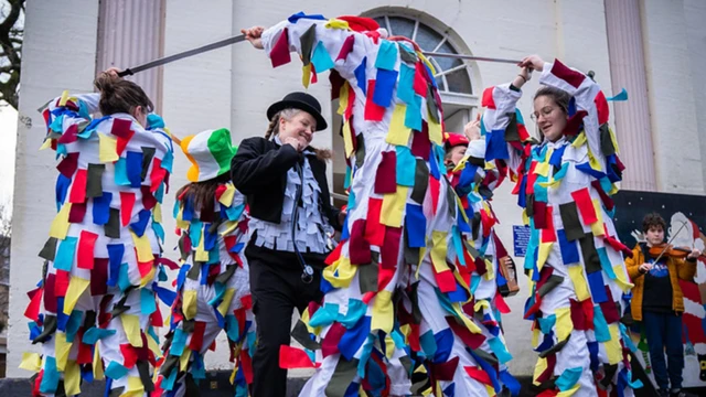 Cinco personas vestidas con trajes blancos decorados con tiras de papel de colores rodean a una dama que baila en el medio y que lleva un bombín, una chaqueta oscura y pantalones.