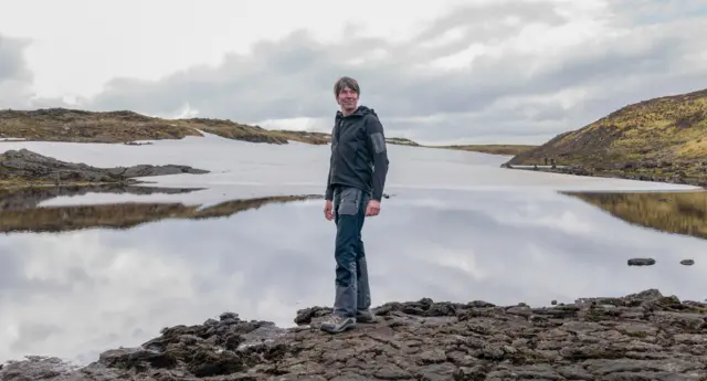 O Prof. Cox apareceaposta eleiçãopé sobre algumas pedras na beiraaposta eleiçãoum grande lago. Colinas são vistas à distância. Ele veste um moletom cinza, calçasaposta eleiçãocaminhada e botas, e sorri