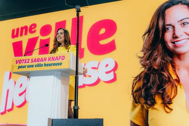 Сара Кнафо на трибуне на фоне собственного портрета на желтом фоне