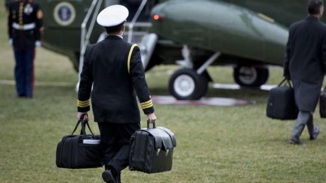 Military officer carries two briefcases to a military aircraft on the White House lawn.