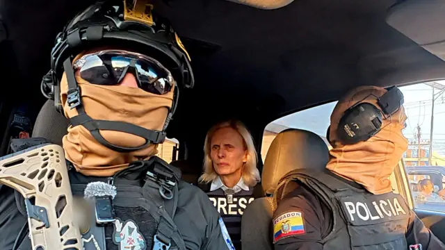 Dos policías ecuatorianos con gafas de sol oscuras, cascos y pasamontañas —que les cubren completamente el rostro— están sentados en un coche frente a Orla Guerin, de la BBC, quien lleva un chaleco antibalas con la palabra "PRENSA" impresa.