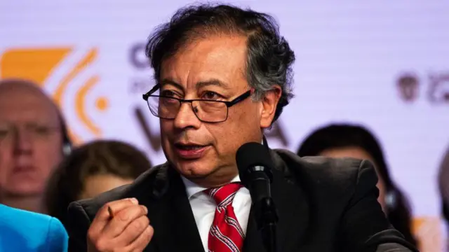 Gustavo Petro, presidente da Colômbia, usa terno preto, camisa branca e gravata vermelha. Ele está de óculos, na frente de um microfone e parece discursar ou falar com pessoas que estão à sua frente. Ao fundo, é possível ver uma série de outras pessoas.