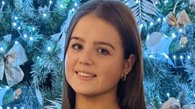 Uma menina de 13 anos está em frente a uma árvore de Natal. Ela veste uma camisa do time de futebol americano Kinawley Brian Borus, nas cores azul-marinho e azul, com o escudo do time no peito. Ela está sorrindo, tem cabelos longos e escuros e usa brincos de argola dourados. A árvore de Natal está decorada com luzes azuis e enfeites dourados claros.