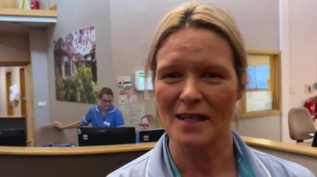 Una fotografía de Nikki Smith vistiendo un uniforme azul, de pie con el personal del hospicio detrás de ella frente a las pantallas de computadora.