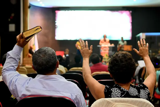 Um homem e uma mulher vistos pelas costas durante culto em igreja evangélica. Os dois estão sentados de frente para o palco com os braços erguidos e ele segura uma bíblia. 
