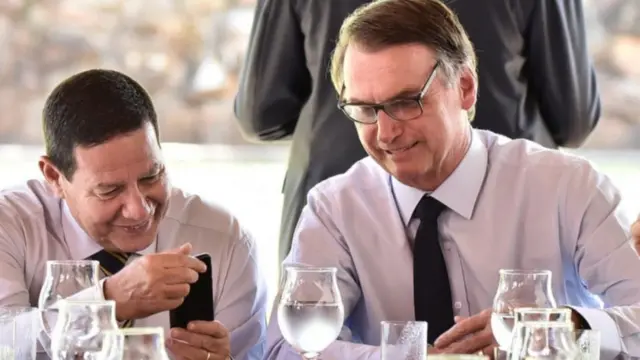 Jair Bolsonaro e Hamilton Mourão em reunião; Bolsonaro mostra algo no celular ao vice presidente e este ri