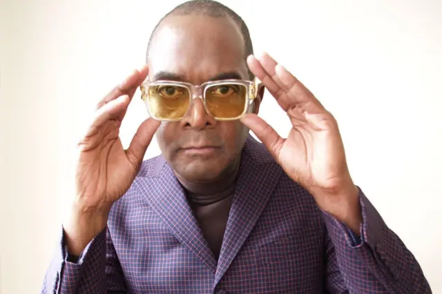 
Dennis Morris akiwa amevalia suti ya blue iliyo na mistari za zambarau na waridi, Amevaa miwani ya njano ambayo ameshikilia na mikono yote miwili