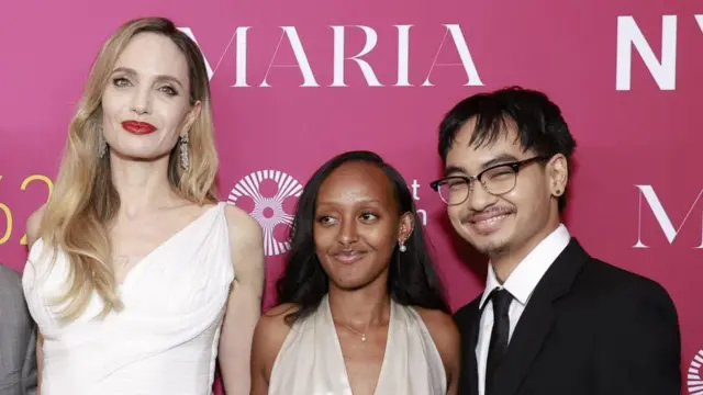 Angelina Jolie, Zahara Marley Jolie oraz Maddox Chivan Jolie-Pitt pozują do zdjęcia przed różową ścianą. Wszyscy są ubrani w eleganckie stroje i uśmiechają się do aparatu.