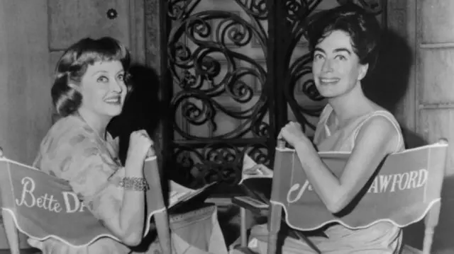 بتی دیویس و جون کرافورد در یک عکس سیاه و سفید، روی صندلی نشسته‌اند به طرف دوربین عکاس برگشته و لبخند می‌زنند