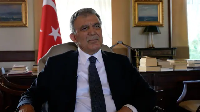 11. Cumhurbaşkanı Abdullah Gül. Arkasında bir çalışma masası ve Türk bayrağı görülüyor. 