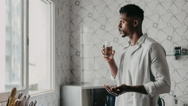 Чоловік тримає склянку з водою і щось у руці і дивиться у вікно