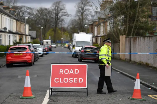 Una calle en Glasgow, Escocia, acordonada con cinta azul y blanca de la policía. Un oficial con la distintiva chaqueta fluorescente habla con alguien que está fuera del encuadre. 