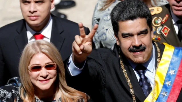 Президент Венесуэлы Николас Мадуро и его жена Силия Флорес приветствуют людей в ходе публичного мероприятия. На нем темные костюм и галстук, желто-сине-красный флаг Венесуэлы надет сверху в виде ленты. Она в темных очках, на ней темное платье с узором