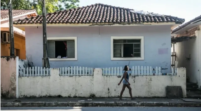 Mulher caminhacalçada à frentecasa azul, com duas janelas e muro