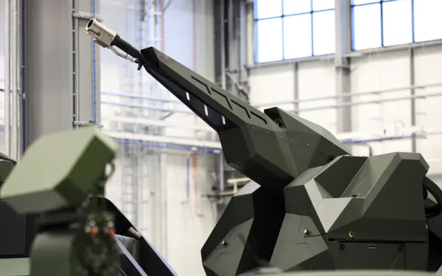 Zbliżenie na górną część dużego, ciemnozielonego pojazdu wojskowego z zamontowaną, podłużną konstrukcją skierowaną ku górze. W tle widoczna jest jasna hala przemysłowa