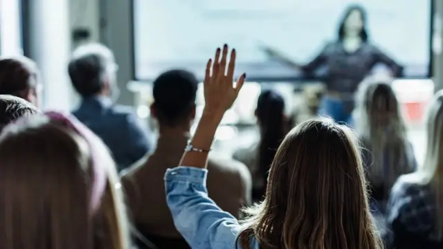 Une femme donne une conférence devant un écran vidéo et une femme dans le public lève la main pour poser une question.