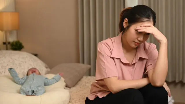 Seorang perempuan dengan baju warna salem duduk di tepi tempat tidur, menutupi mata dengan sebelah tangannya, sementara di belakangnya seorang bayi dengan baju terusan warna biru menangis.