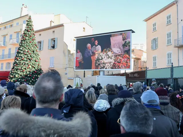 Толпа стоит перед большим экраном, на котором показывают церемонию прощания