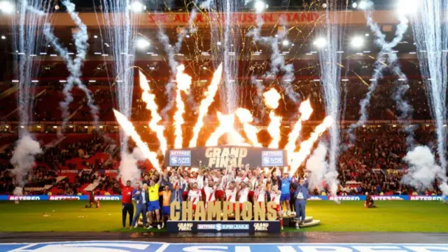 Hull KR celebra su victoria en la Gran Final de la Superliga: todo el equipo anima frente a las gradas mientras levanta el trofeo mientras se escuchan fuegos artificiales de fondo.