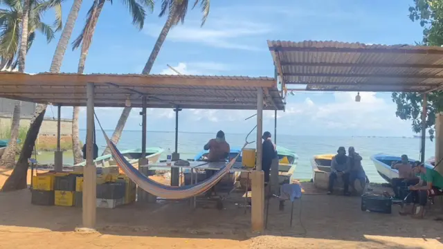 Al fondo se ve la playa en cuya orilla hay varios botes de pescadores. Más adelante hay una especie de estación rudimentaria con un techo de metal bajo el cual hay unas mesas rústicas y unas cestas plásticas de colores. Allí entregan los pescadores sus cangrejos para ser pesados antes de ser enviados a procesar.