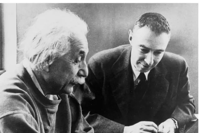 Oppenheimer e Einstein sorrindo contidamente e olhando para baixo, na mesma direção