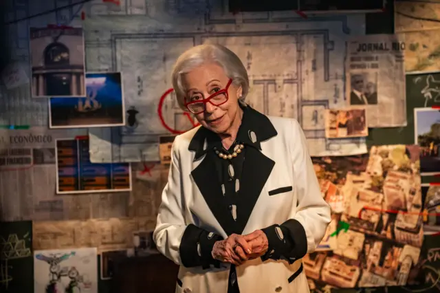 Fernanda Montenegro, de cabelos brancos e usando óculos de armação vermelha, aparece em primeiro plano diante de um painel cheio de fotos, recortes de jornal e um mapa com marcações em vermelho, como em uma parede de investigação. Ela veste um blazer branco com detalhes pretos, camisa escura com estampa clara e um colar de pérolas, mantendo as mãos juntas à frente do corpo e olhando para a câmera com expressão inquisitiva.