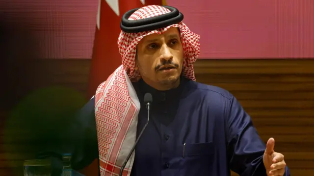 Mohammed bin Abdulrahman bin Jassim al-Thani gesticulando ao falar no microfone, em frente a bandeira 