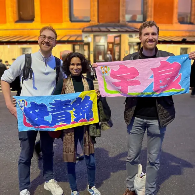 ロイヤル・アルバート・ホールの前でアフリカ系の女性1人と白人男性2人が、青で「安青錦」、ピンクで「宇良」と書かれた鮮やかな力士タオルを広げてカメラに向かって笑っている