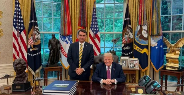 Foto mostrando Donald Trump dentro do salão oval da Casa Branca, com bandeira dos Estados Unidos ao fundo. Flávio está em pé e Trump sentado