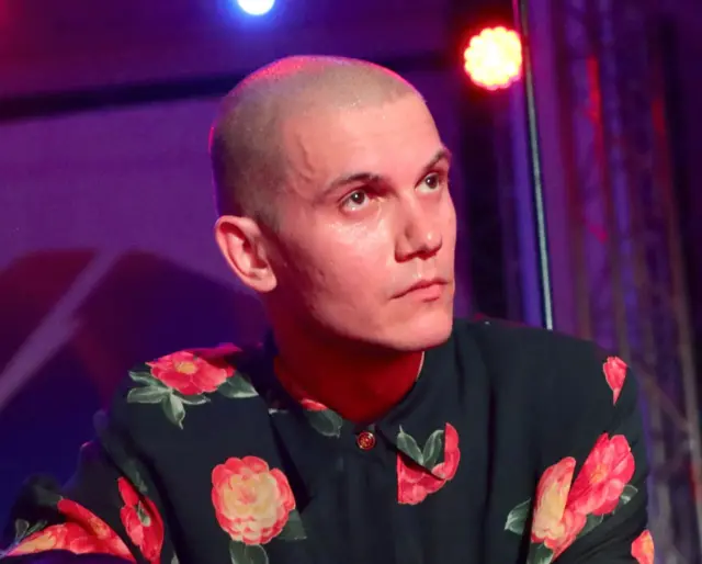 Николай Комягин в черной рубашке с красными цветами, наголо обритый, смотрит куда-то вверх. Задний план сиреневого цвета