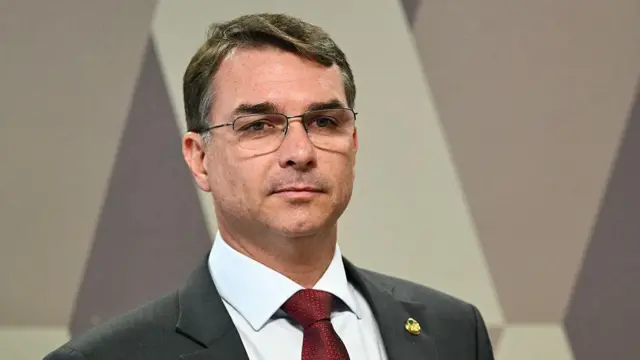 Flavio Bolsonaro é um homem branco. Ele usa óculos e está com um terno cinza e gravata vermelha.