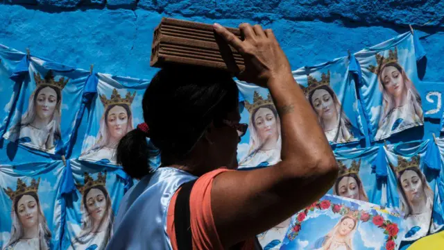 Uma mulher carrega um tijolo em cima da cabeça diante de um muro azul coberto por estampas repetidas de uma figura religiosa.