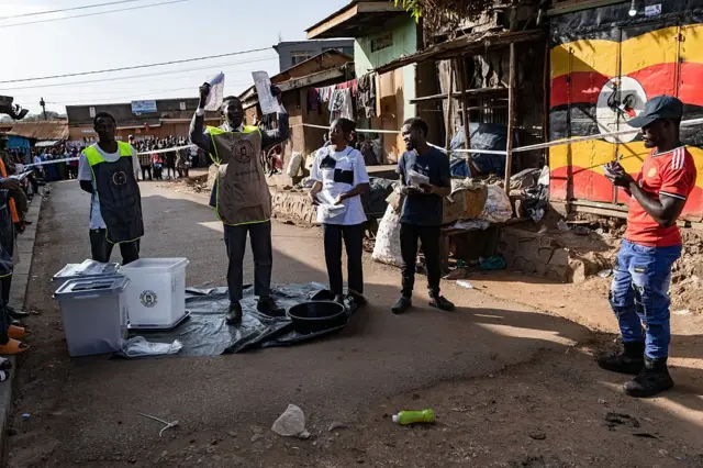 Посреди трущоб на ровном участке асфальта расстелен пластик, на нем стоит избирательный бюллетень. Рядом с ним стоят пять человек, трое в обычной одежде, на двоих надеты длинные жилетки с надписями, эмблемами и светоотражающими элементами. Чуть поодаль натянута лента, за ней столпились люди.