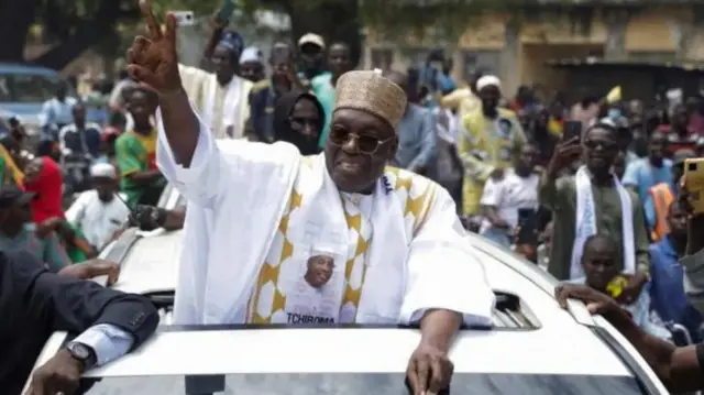 Issa Tchiroma Bakary wear one white traditional attire, e stand inside one motor and raise one of im hand up. Im supporters surround am.