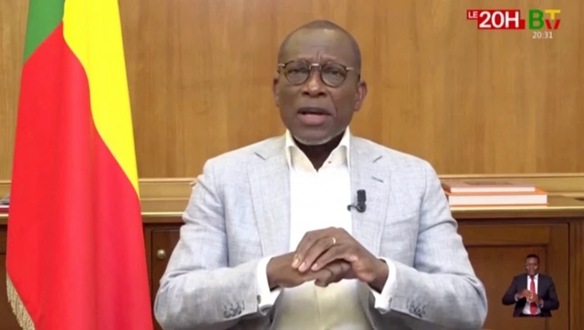 Benin Cumhurbaşkanı Patrice Talon kravatsız şekilde, beyaz bir gömlek gri ceket giymiş, yanında ülke bayrağı var. Konuşurken sakin görünüyor