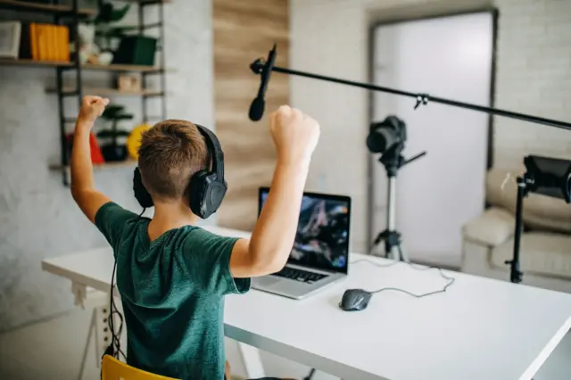 Menino de costas gravando vídeo com equipamentos de filmagem e laptop; ele está sentado e erguendo os braços
