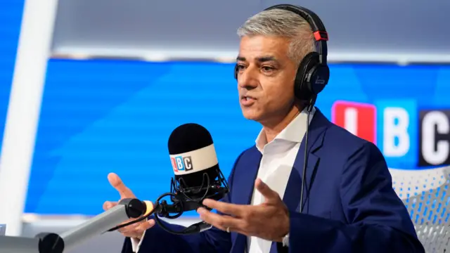 Sadiq Khan speaking in the LBC studio. He wears a white shirt, navy blue suit jacket, and headset. He is gesturing with his arms open.
