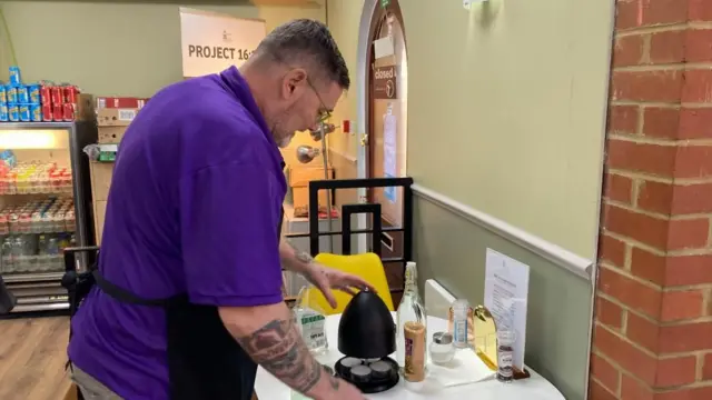 Stan trabaja en la cafetería, inclinado y limpiando una mesa con objetos encima. Está de lado a la cámara con un delantal y una blusa morada. Tiene tatuajes en el antebrazo derecho.